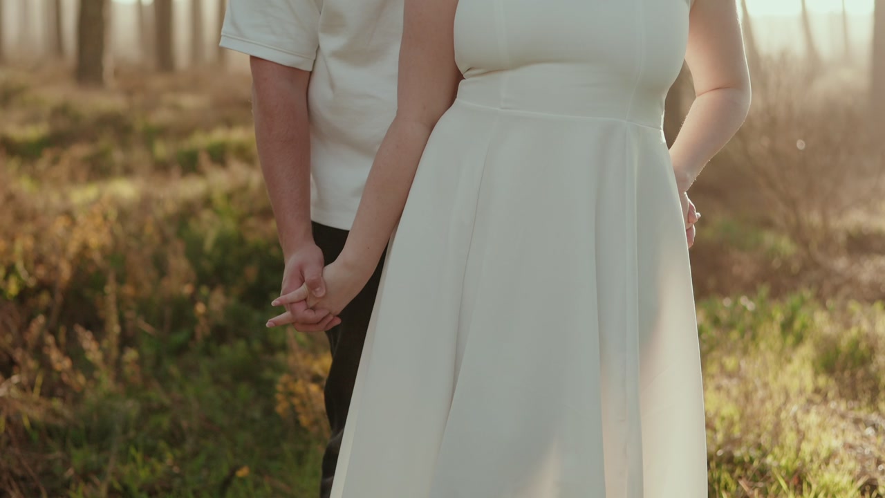 Close view of bride in white dress holding hands with partner standing in forest with warm sunlight