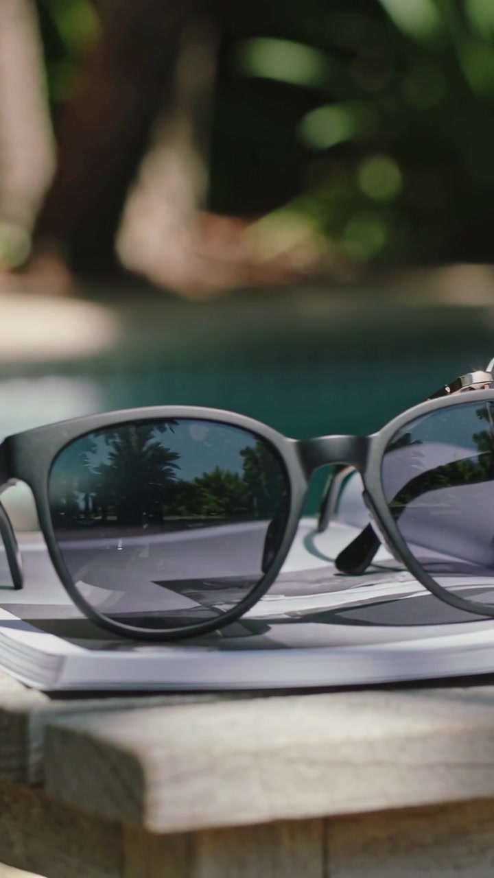 Vertical video: Panning camera revealing two sunglasses on wood table by pool, reflecting palms
