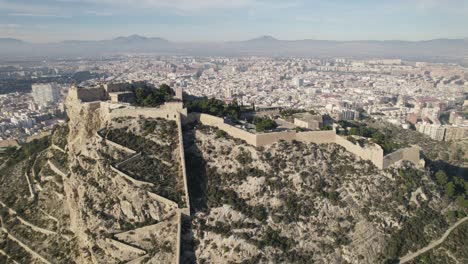 Fortaleza-De-Santa-Bárbara-En-La-Cima-Del-Monte-Benacantil,-Fondo-Del-Paisaje-Urbano-De-Alicante,-Vista-Aérea-En-órbita