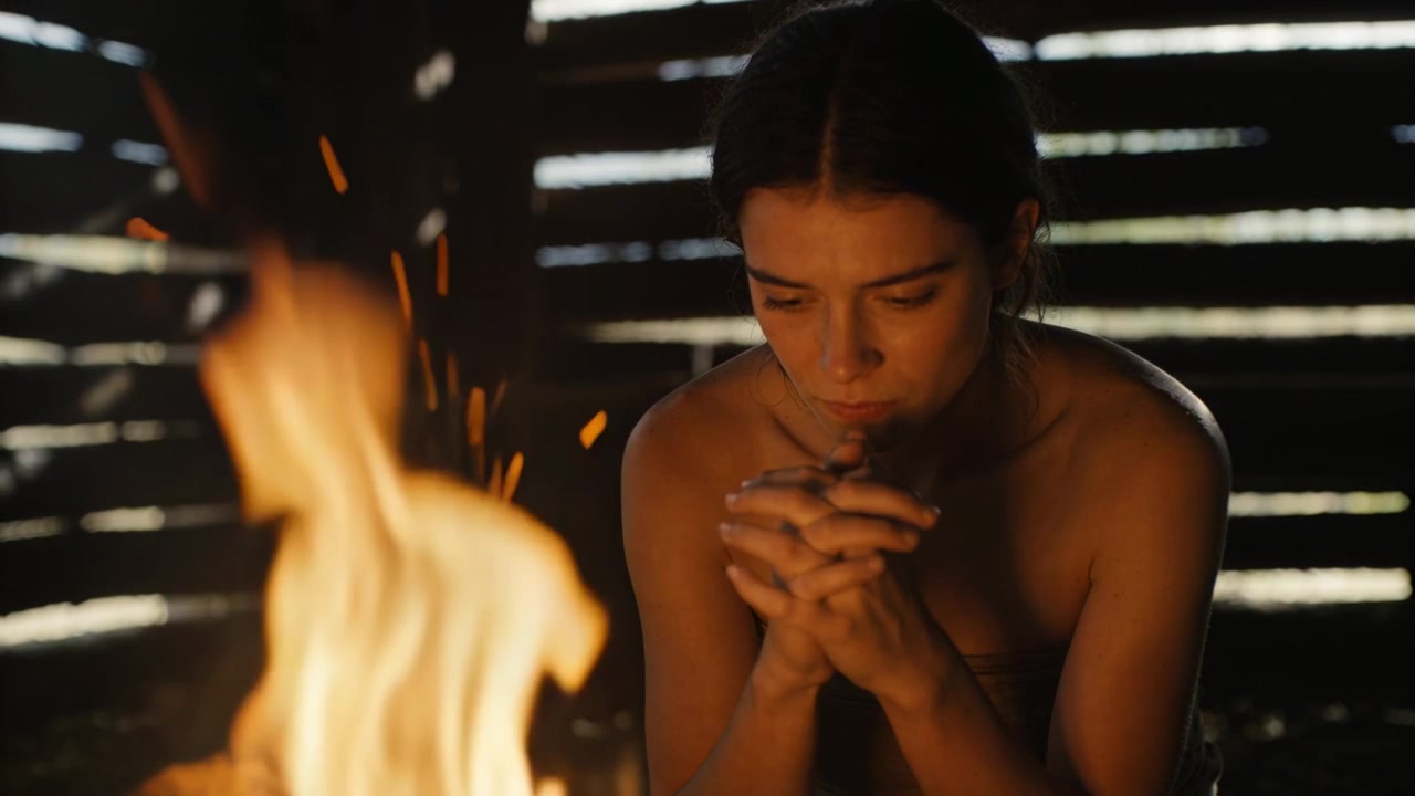 Chamas a arder, mulher de ombros nus com as mãos em concha, soprando em direção a uma fogueira de madeira numa cabana, aquecendo as mãos.
