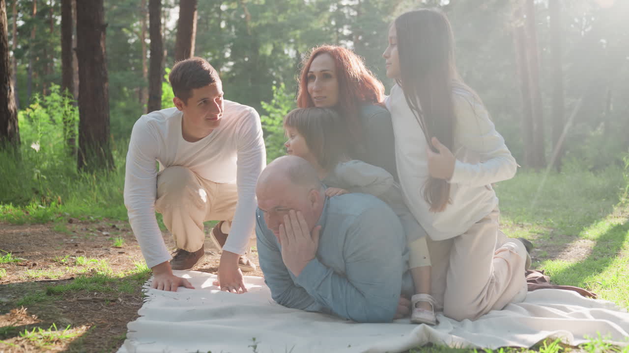 Dad lying comfortably on picnic mat while wife and daughters rest on his back surrounded by sunlight and forest greenery, creating warm family bonding moment filled with love