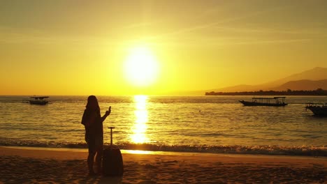 Silueta-De-Una-Mujer-Joven-Con-Un-Equipaje-Tomando-Selfies-En-La-Playa-De-Arena-Paradisíaca-Durante-La-Puesta-De-Sol-Dorada