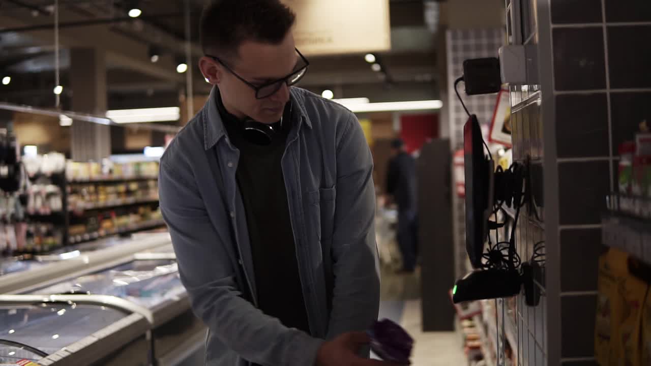 Cheerful man in glasses and headphones on neck pusing his trolley full of food while shopping at the local supermarket. Scanning the pack with a special scanning equipment to get know the cost. Slow motion