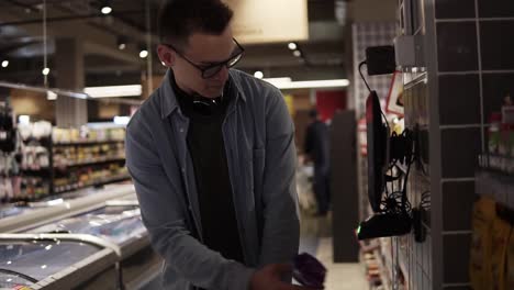 cheerful man in glasses and headphones on neck pusing his trolley full of food while shopping at the local supermarket. scanning the pack with a special scanning equipment to get know the cost. slow motion