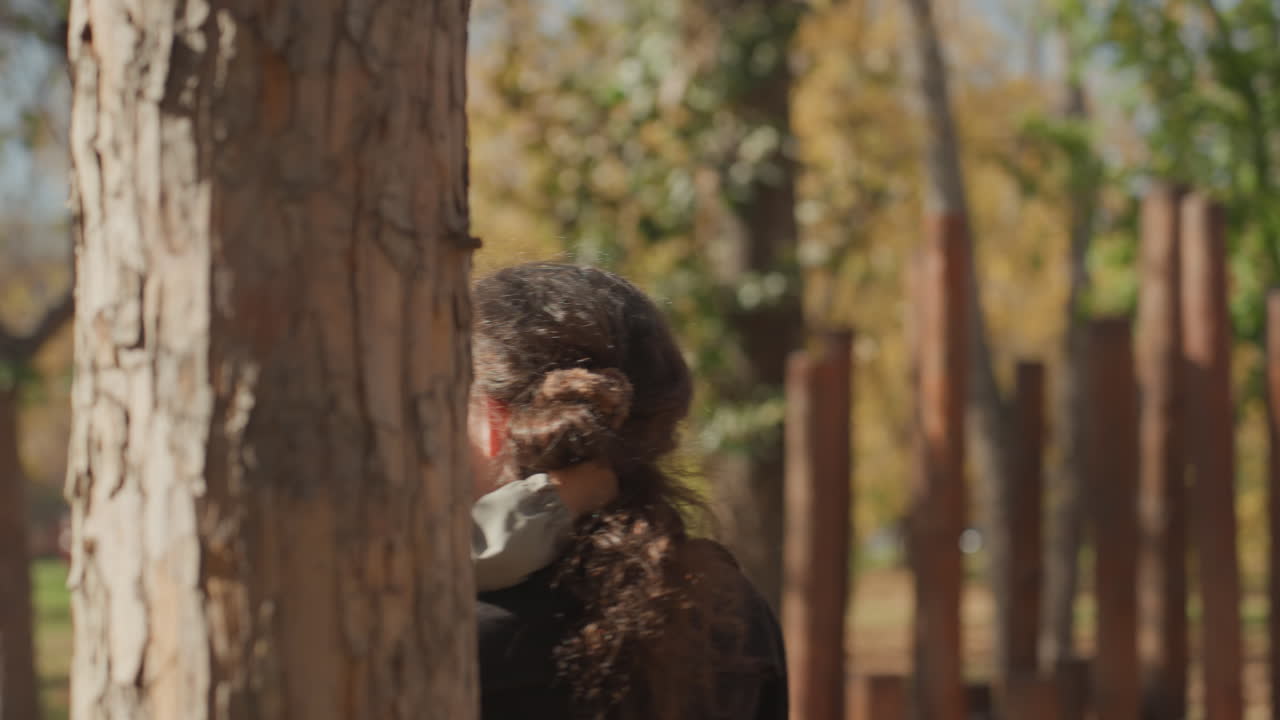 Madre che tiene in braccio il bambino piccolo dietro un albero nel parco, un momento spontaneo di cucù con la luce dorata autunnale, pali di legno e foglie cadute, bambino sorridente in giacca, abbraccio affettuoso, una calda scena genitoriale