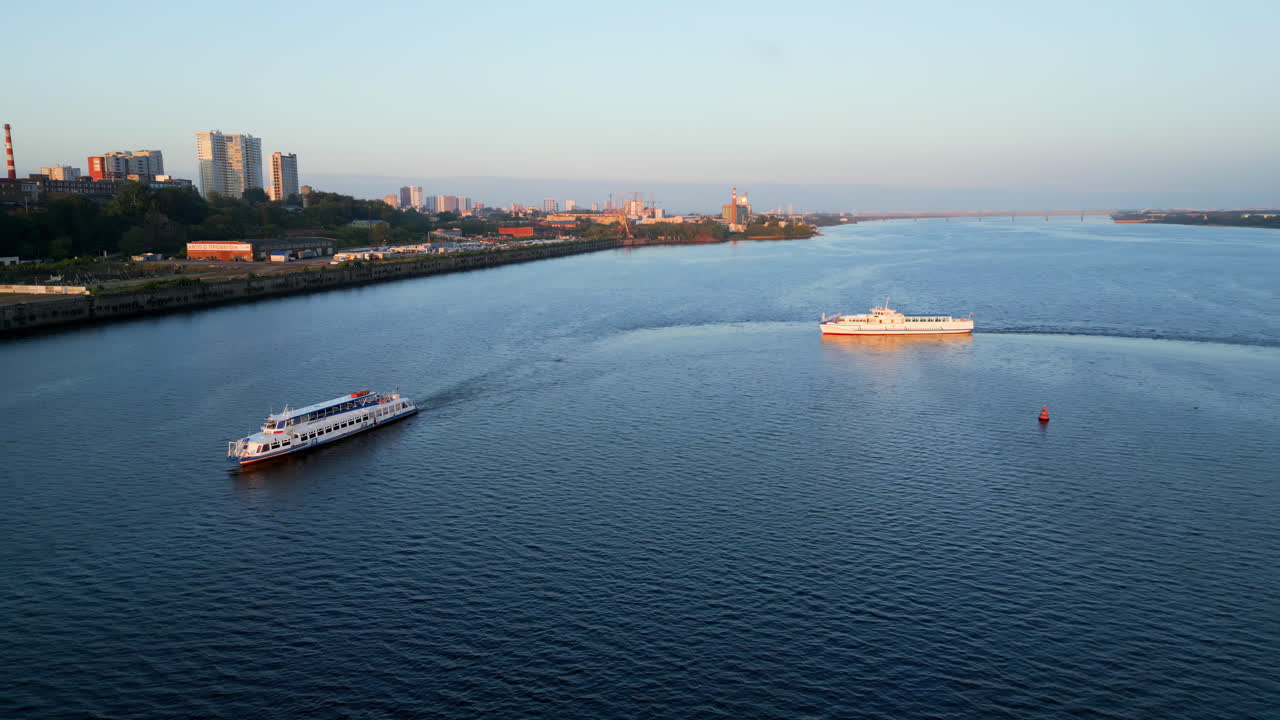 Two passenger boats navigating a wide river with a city skyline and industrial buildings on the shore