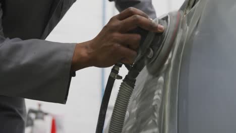 African-American-male-car-mechanic-wearing-a-face-mask-and-polishing-a-side-of-a-car-with-a-grinder