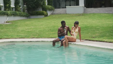 Happy-diverse-couple-wearing-swimming-suits-and-drinking-drinks-at-swimming-pool-in-garden