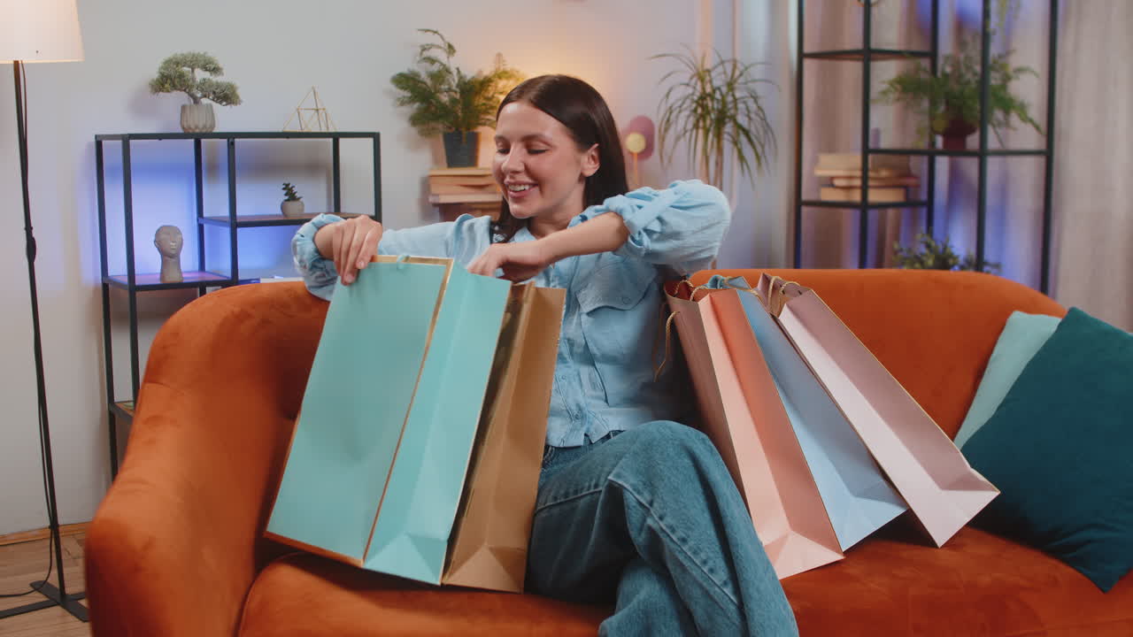 joven feliz caucásica adicta a las compras mujer con bolsas de compras sentada en el sofá en la sala de estar en casa