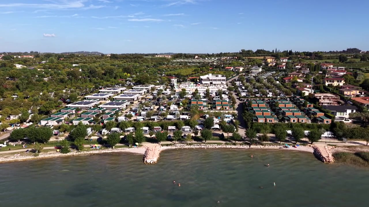 Bungalós de tejado verde bordean un camping denso junto a una costa curva, un pequeño embarcadero de piedra se extiende hacia aguas tranquilas de color turquesa marcadas por boyas naranjas cerca del lago de Garda, Italia. Toma aérea lateral de gran angular con dron