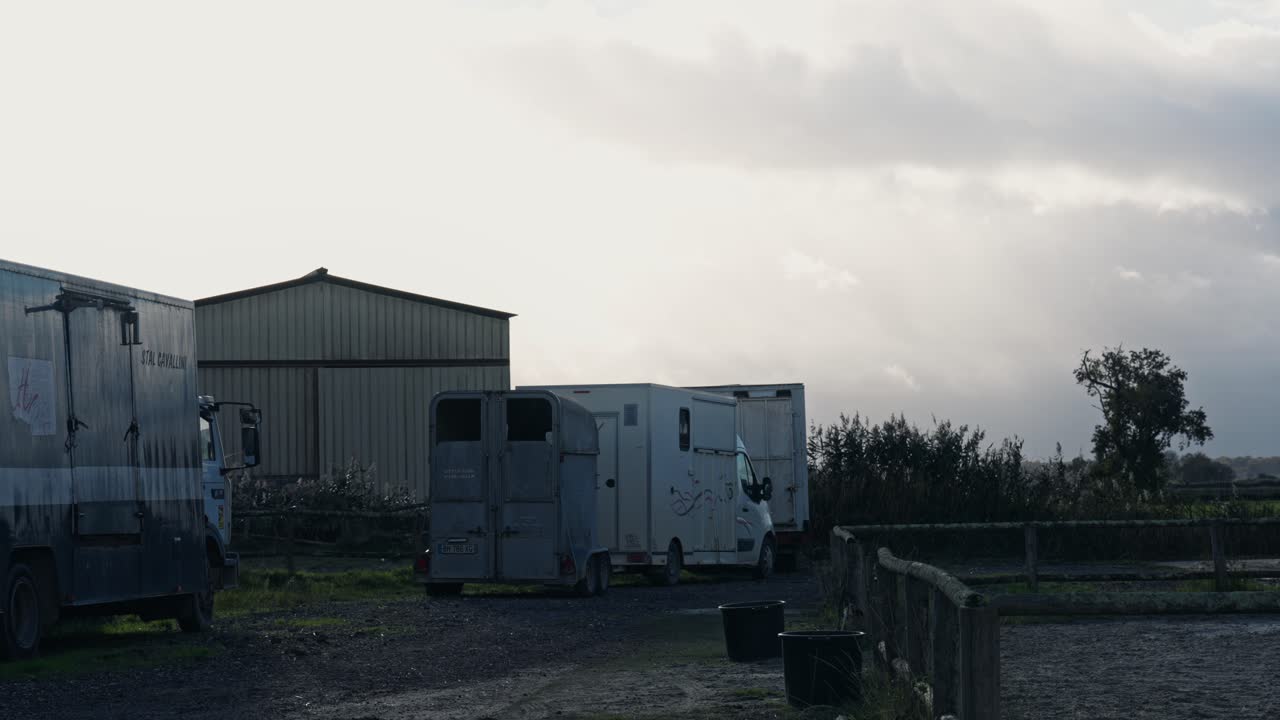 Una fila di rimorchi bianchi per cavalli e camion da trasporto parcheggiati vicino a un grande capannone metallico. Attrezzatura professionale per il trasporto equestre in un ambiente rurale di campagna sotto un cielo nuvoloso