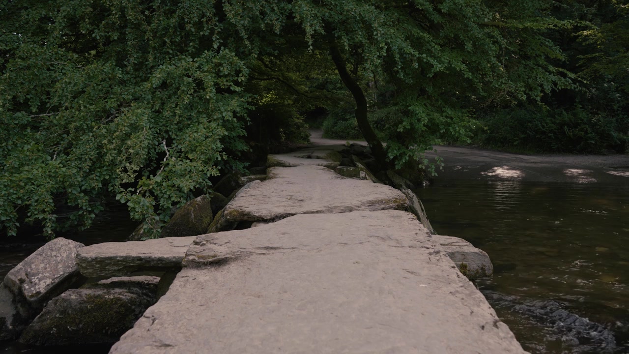 Tarr Steps showing the iconic clapper bridge stretching across the River Barle. The scene emphasises stillness and natural beauty, subtle movement in the water and leaves enhancing the peaceful scene.