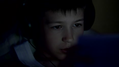 boy plays on the tablet lying in the dark in headphones