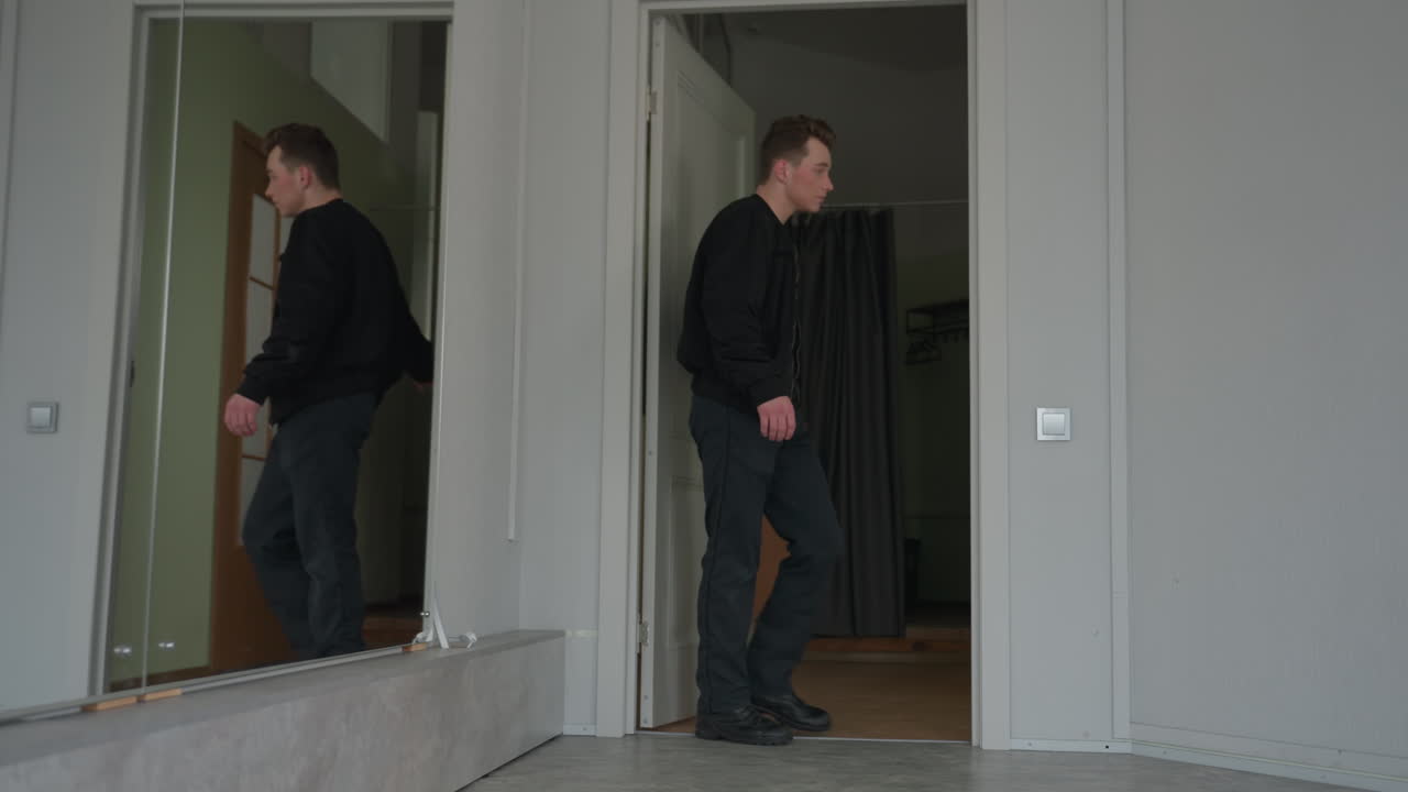 Young man closing apartment door after entering room, captured from behind in black jacket, mirror on wall reflecting surrounding corridor, soft daylight illuminating minimalist interior