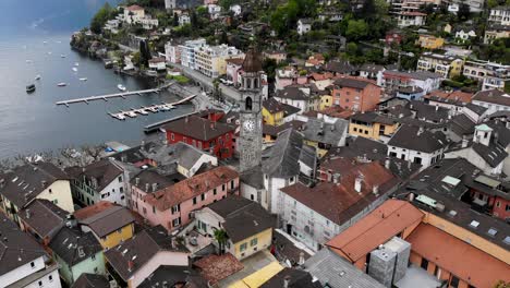 Sobrevuelo-Aéreo-Sobre-Los-Tejados-De-Ascona,-Suiza,-Con-Vistas-A-La-Torre-De-La-Iglesia-Y-A-Las-Orillas-Del-Lago-Maggiore,-Lleno-De-Barcos-Y-Corwds-En-El-Paseo-Marítimo