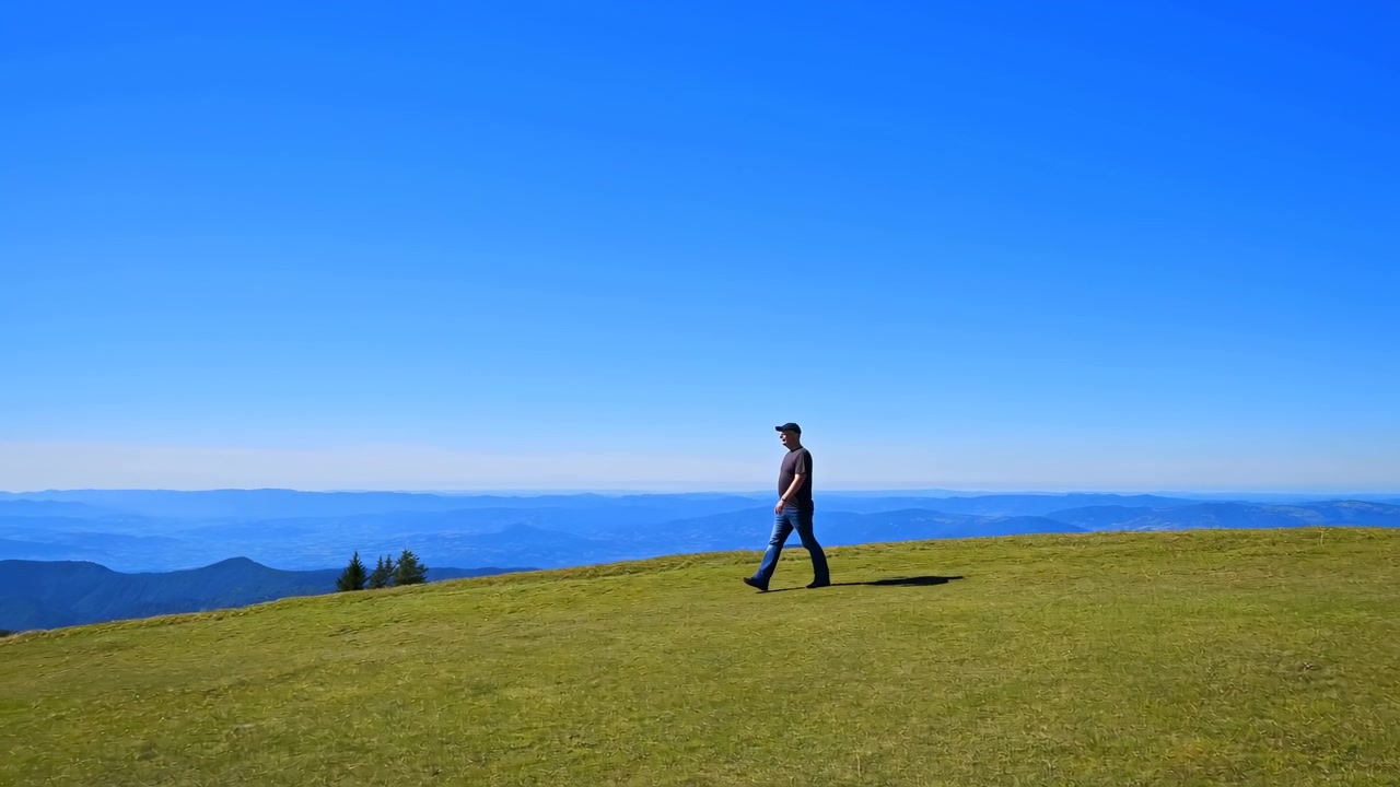 Single man walking along grassy ridge with lifted step and slight forward lean, set against layered mountains, blue sky, bright midday light, short shadow, and wide open landscape, with walking, man