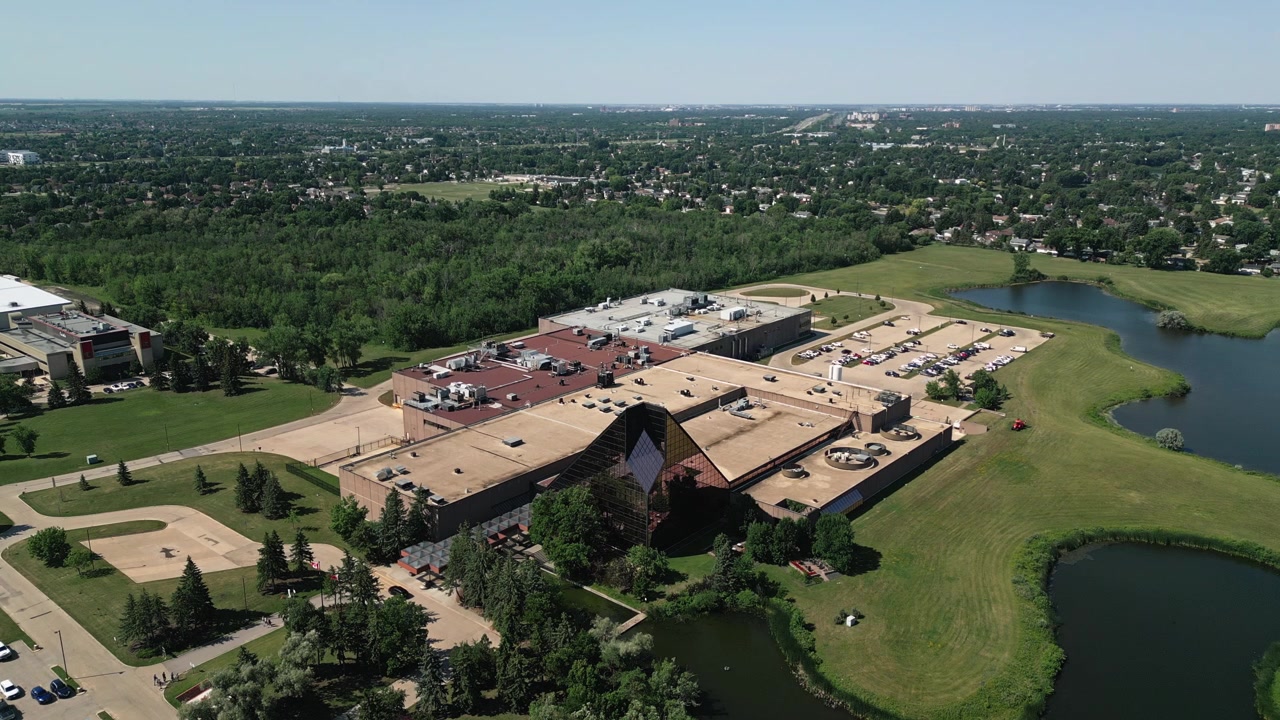 Ripresa aerea statica con drone del complesso della Zecca Reale Canadese con alberi circostanti, quartieri residenziali e lo skyline di Winnipeg in una luminosa giornata estiva