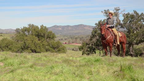 el vaquero se sienta en su caballo en una colina californiana verde con su granero en la distancia