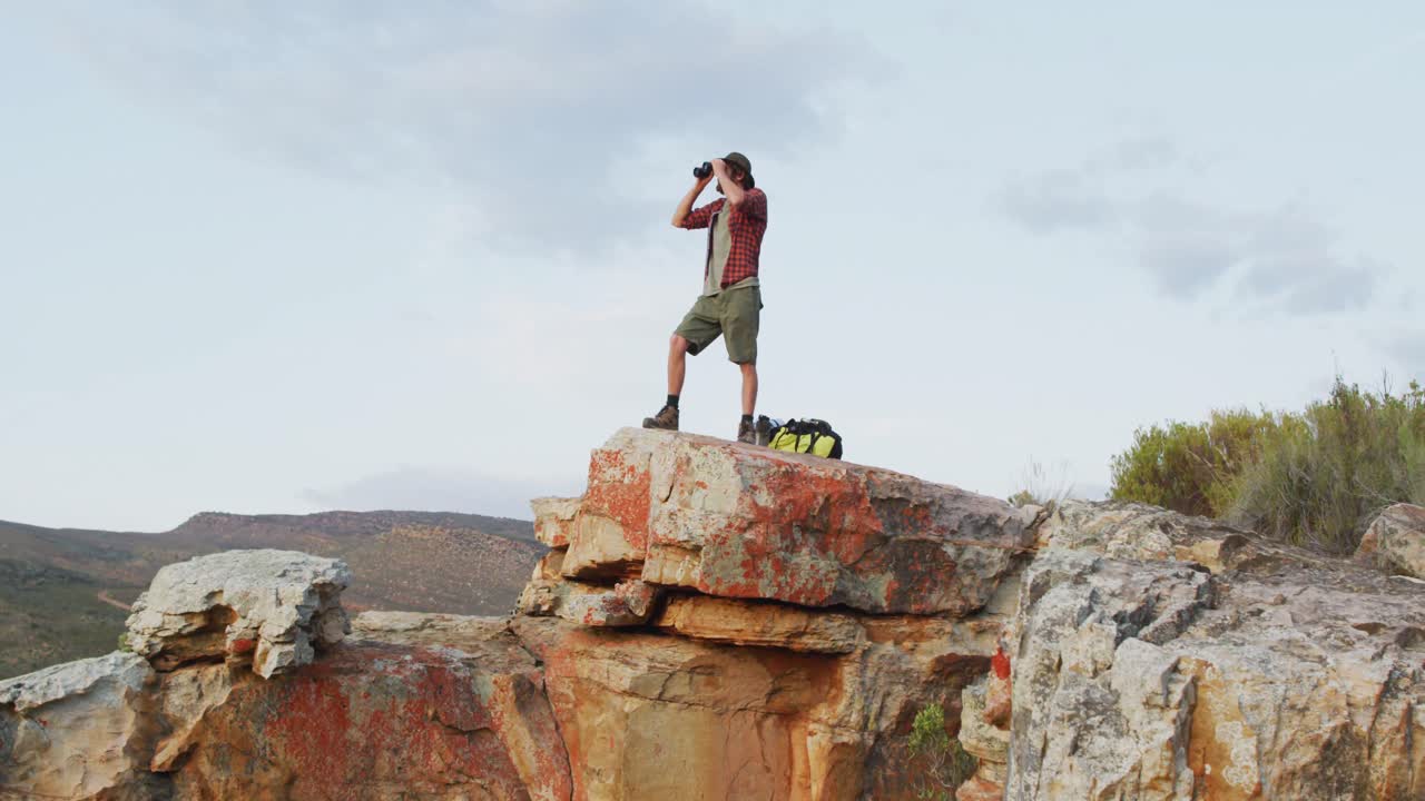 sobreviviente caucásico masculino usando binoculares, de pie en el pico de la montaña en el desierto