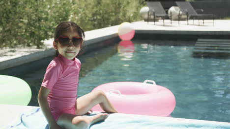 Chica-Birracial-Con-Traje-De-Baño-Rosa-Y-Gafas-De-Sol-En-Forma-De-Corazón-Sentada-Junto-A-Una-Piscina