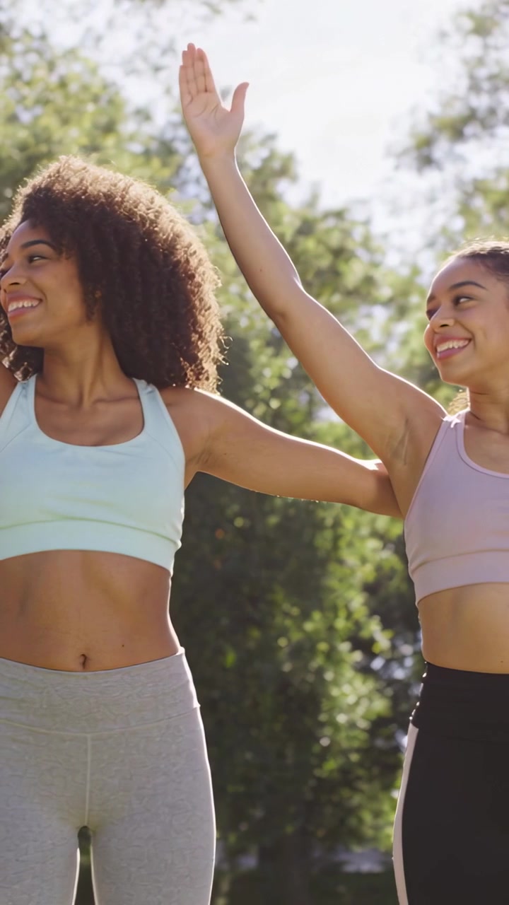 Amiche di diverse etnie che imitano i sollevamenti delle braccia in un parco soleggiato, indossando reggiseni sportivi e leggings