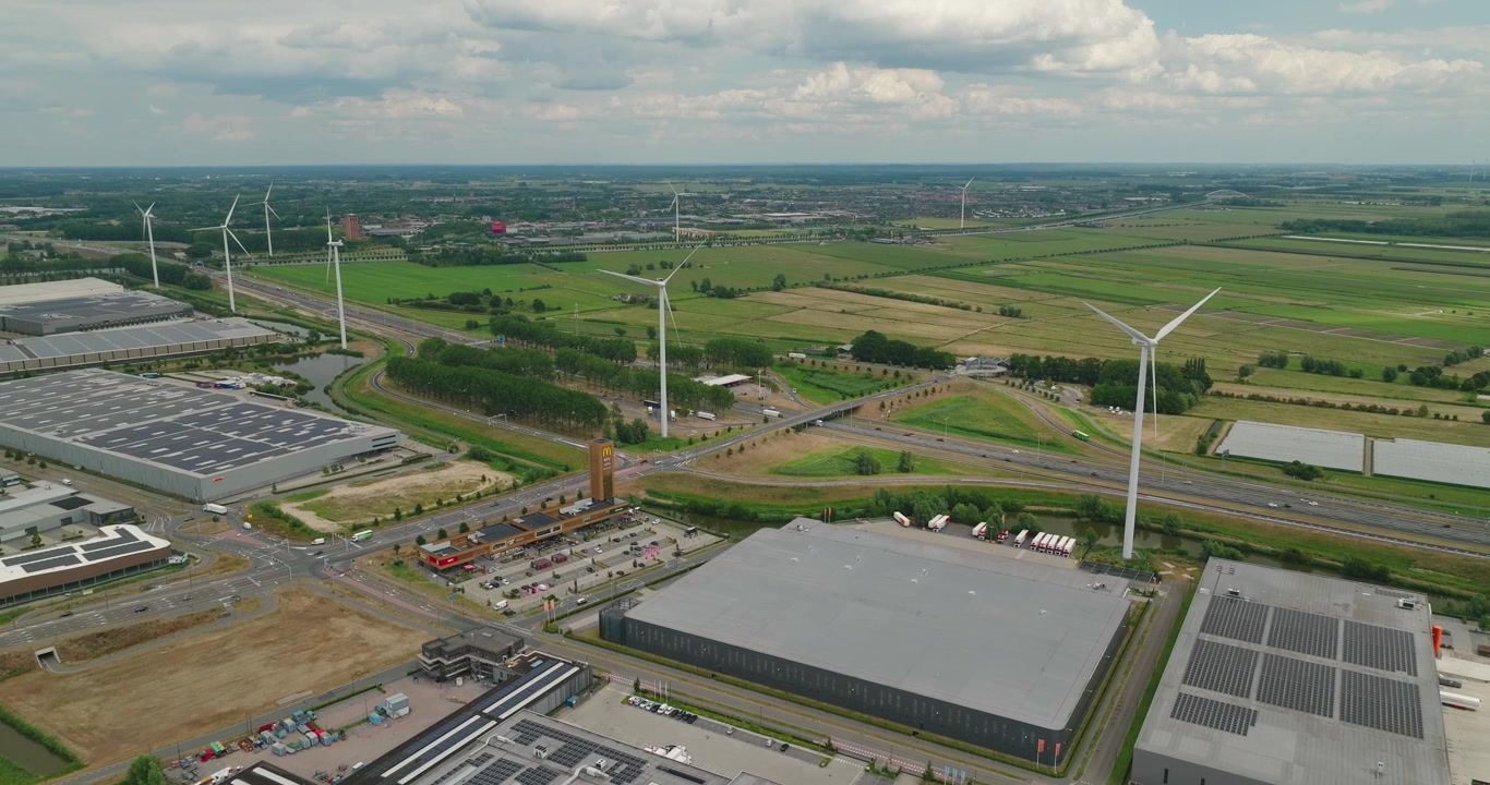Drone flies over a modern business park with large warehouses, windmills and a flat Dutch countryside landscape stretching across the horizon.
