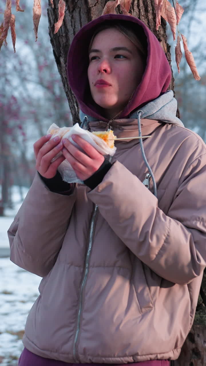 giovane ragazza in abito invernale che si riposa sull'albero che mangia spuntini nel parco all'aperto durante l'inverno, riflesso di luce dallo sfondo, che si gode il momento del cibo nel parco, le vibrazioni del tempo freddo