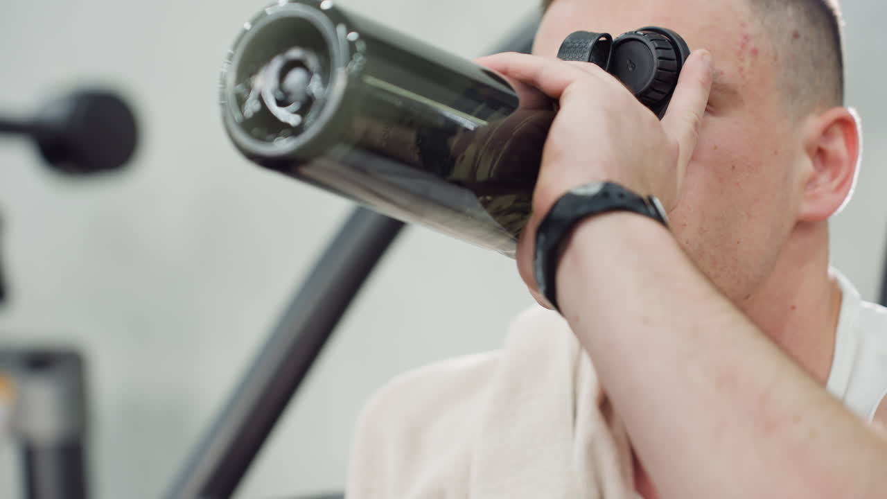 Close up of fitness fanatic drinking water during break with relief on face, seated on gym bench, arm raised gripping bottle firmly, sweat visible under gym lights after intense workout