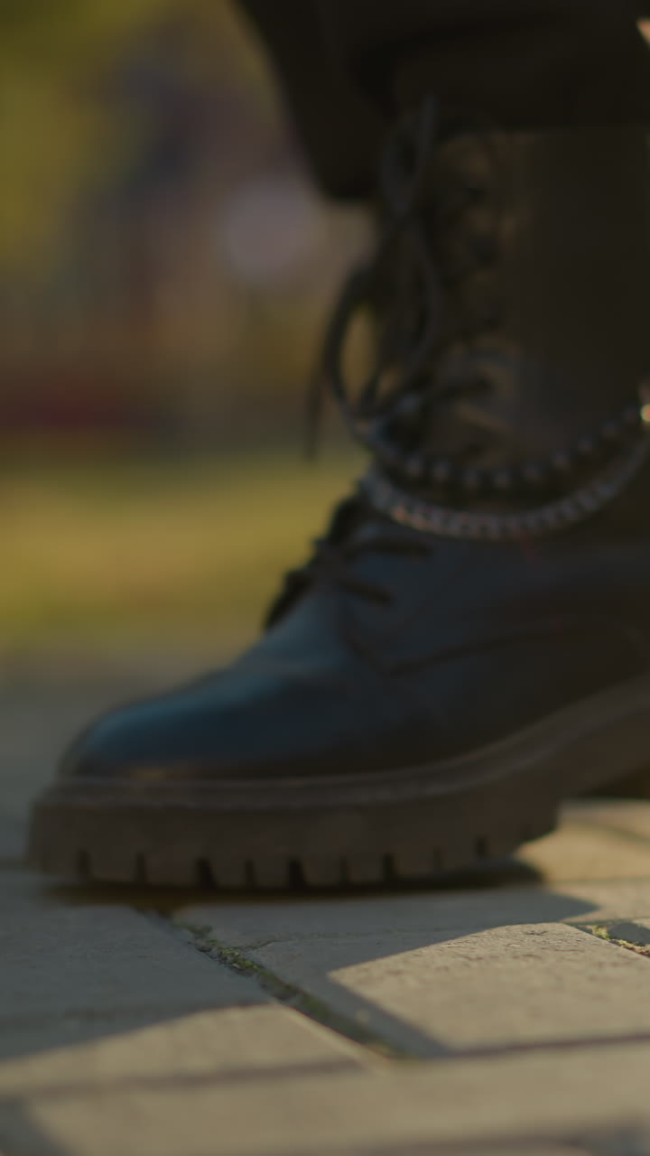 A close-up shot of black boots stepping on a paved path in a sunlit park. The focus is on the boots, showing the details of the laces and sturdy soles. The background is softly blurred