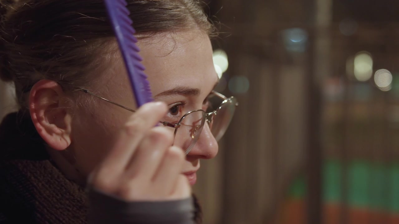 Donna caucasica all'aperto di notte che si pettina, pettine viola attraverso lo chignon mentre sistema la catenella degli occhiali, cappotto marrone caldo e sciarpa spessa, sorriso gentile con bokeh di luci sfocate della città, momento spontaneo di cura di sé.