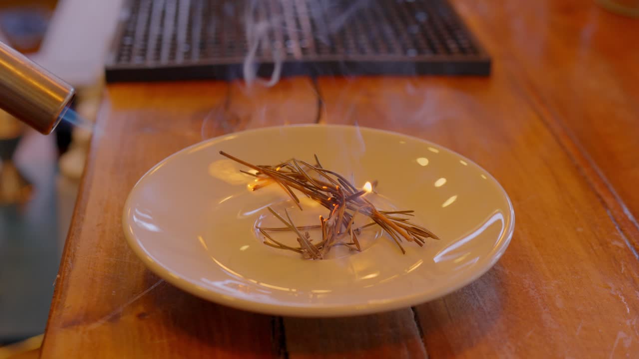preparación de una bebida, se ve que un soplete está quemando hojas de romero encapsuladas por una copa de vidrio para capturar la esencia del olor del romero