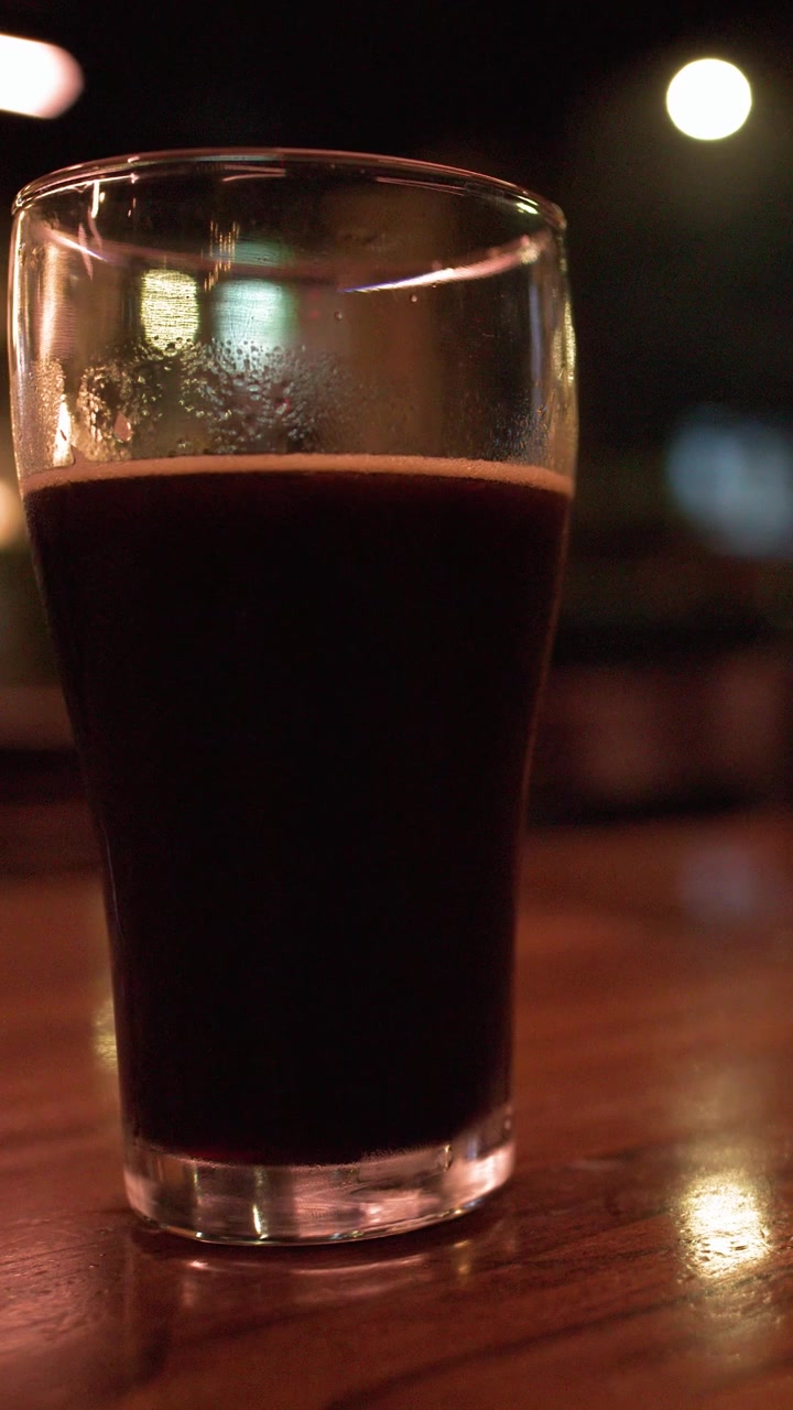 A pint of dark stout beer rests on a wooden table in a dim bar