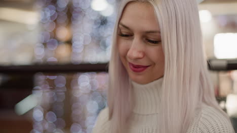 portrait of beautiful lady seated on bench in mall tapping smartphone with gentle smile under decorative bokeh lights beside glass railing festive indoor ambiance with stylish nails and chic watch