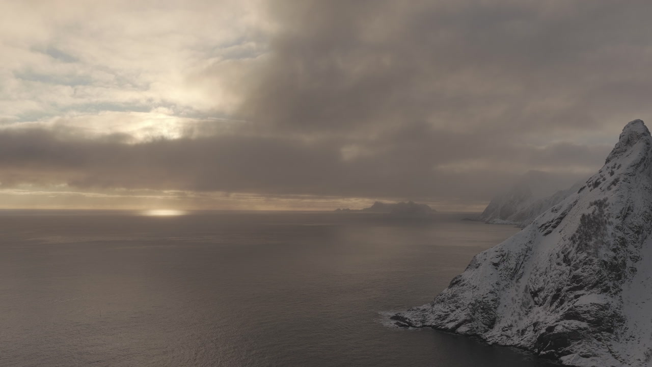 A breathtaking aerial view of the sun setting behind thick clouds over the arctic ocean and snowcovered peaks in Lofoten, Norway.