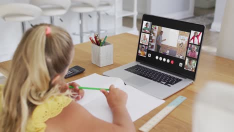 Schoolgirl-using-laptop-for-online-lesson-at-home,-with-diverse-teacher-and-class-on-screen