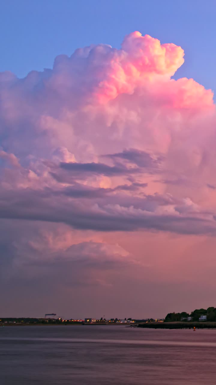 Timelapse shows towering pink-lit cumulonimbus over riverside skyline, flickering with intermittent lightning strikes