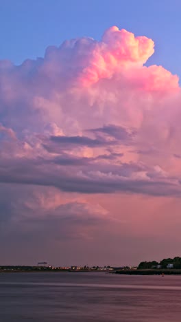 timelapse shows towering pink-lit cumulonimbus over riverside skyline, flickering with intermittent lightning strikes