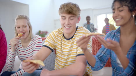 un grupo feliz y diverso de amigos adolescentes sentados en el sofá y comiendo pizza en casa, en cámara lenta