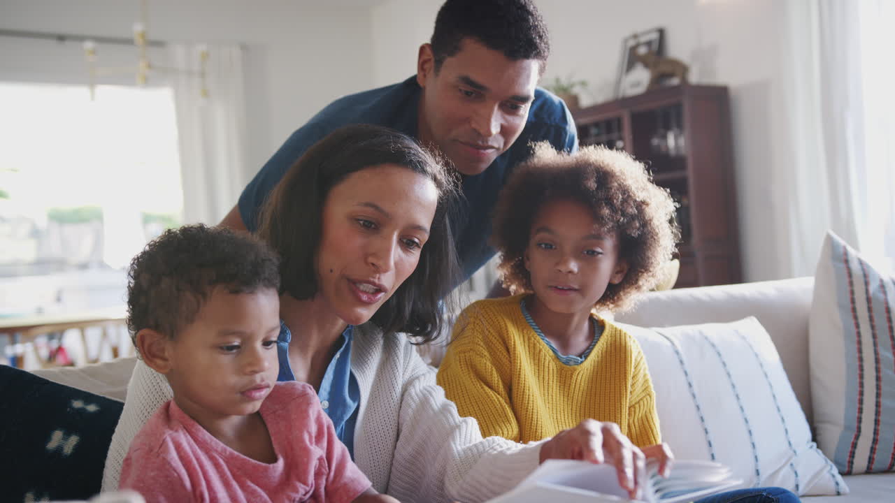 Giovane madre afroamericana che legge un libro ai suoi due figli, il papà che osserva alle loro spalle, primo piano.