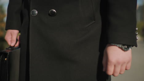 close-up of individual wearing chain wristwatch and black coat while strolling outdoors holding briefcase, background features blurred greenery, modern urban landscape, and a moving car under