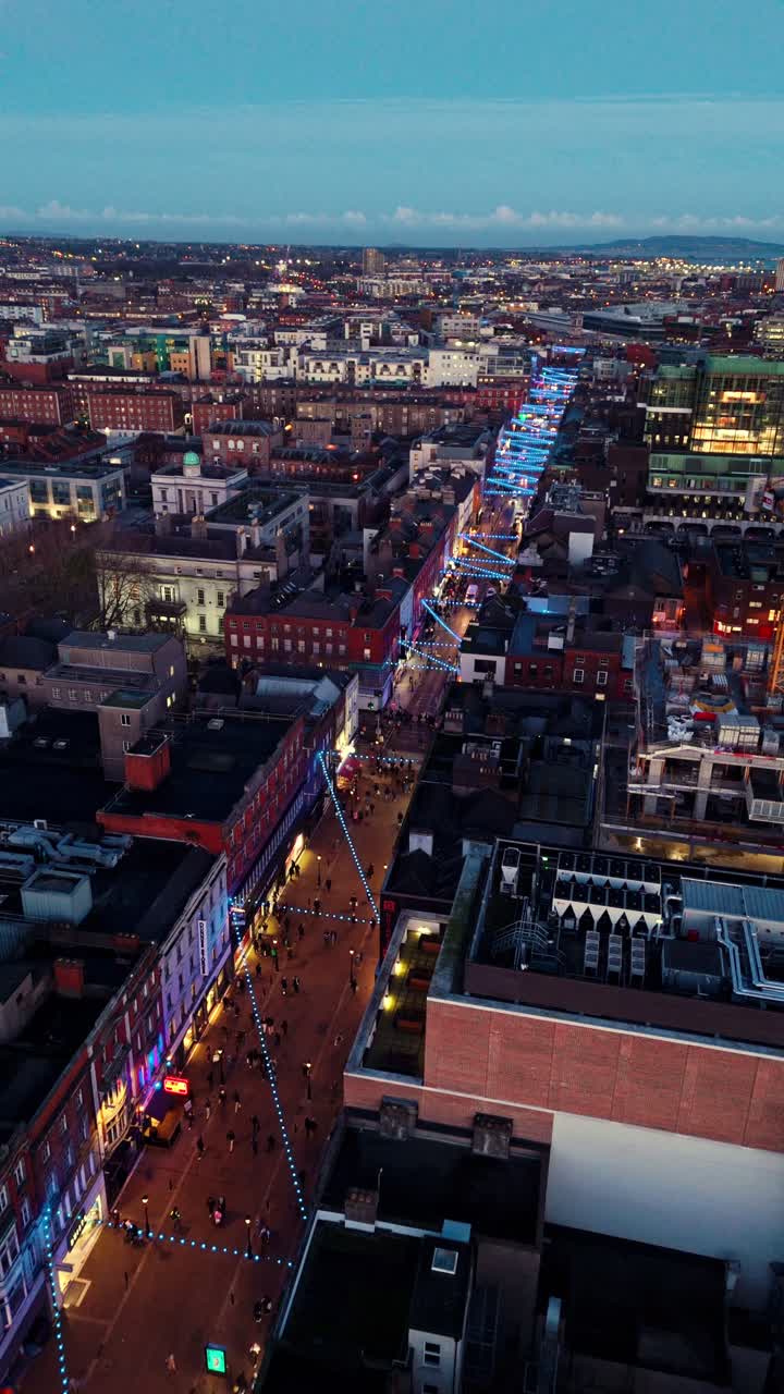 portrait panning drone shot over O'Connell street in Dublin at twilight blue hour with lit street in winter and cityscape with Irish buildings and tourists walking below of the busy street