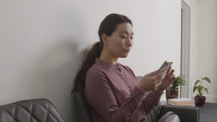 Male And Female Candidates In Office Waiting For Job Interview Looking At Mobile Phone And Reading Notes