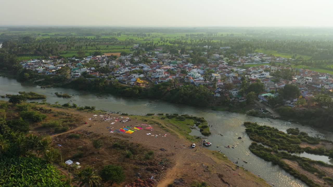 Vista aerea di un villaggio sul fiume che cattura la vita rurale quotidiana, con gli abitanti del villaggio che lavano i vestiti e i veicoli vicino a un fiume calmo, con case, campi verdi e uno stile di vita rurale tranquillo alla luce del giorno