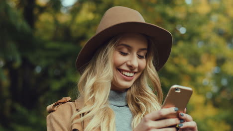 Close-up-view-of-caucasian-young-blonde-woman-wearing-a-hat-and-texting-on-smartphone-in-the-park-in-autumn