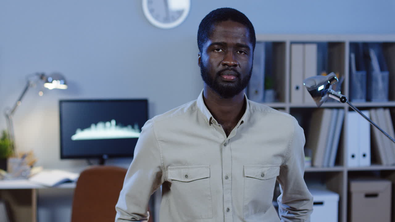 joven oficinista sonriendo a la cámara y mostrando un gesto bien con una mano en la oficina por la noche