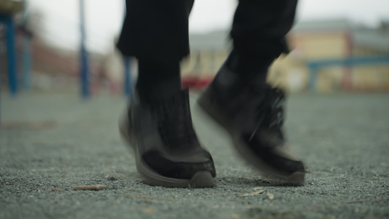 Close-up of a person s legs jogging in an increasing pace, in black jeans and black sneakers, with a blurred background that includes a bench and a painted yellow building