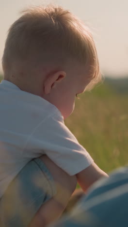 una vista trasera de un niño pequeño con una bufanda en un campo cubierto de hierba, inclinado hacia adelante mientras juega. un fondo borroso presenta un vestido azul fluido y un niño parcialmente visible acostado cerca