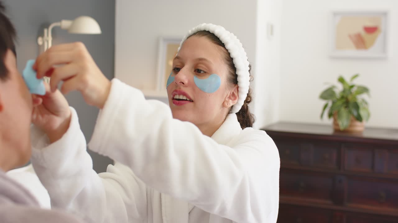 Coppia multietnica a casa, donna in accappatoio bianco che offre e applica una maschera blu per il contorno occhi per la cura della pelle.