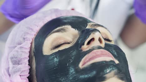 cosmetician in purple gloves carefully puts a young woman on the face purifying black mask. scin care, beautician applying face mask cream.