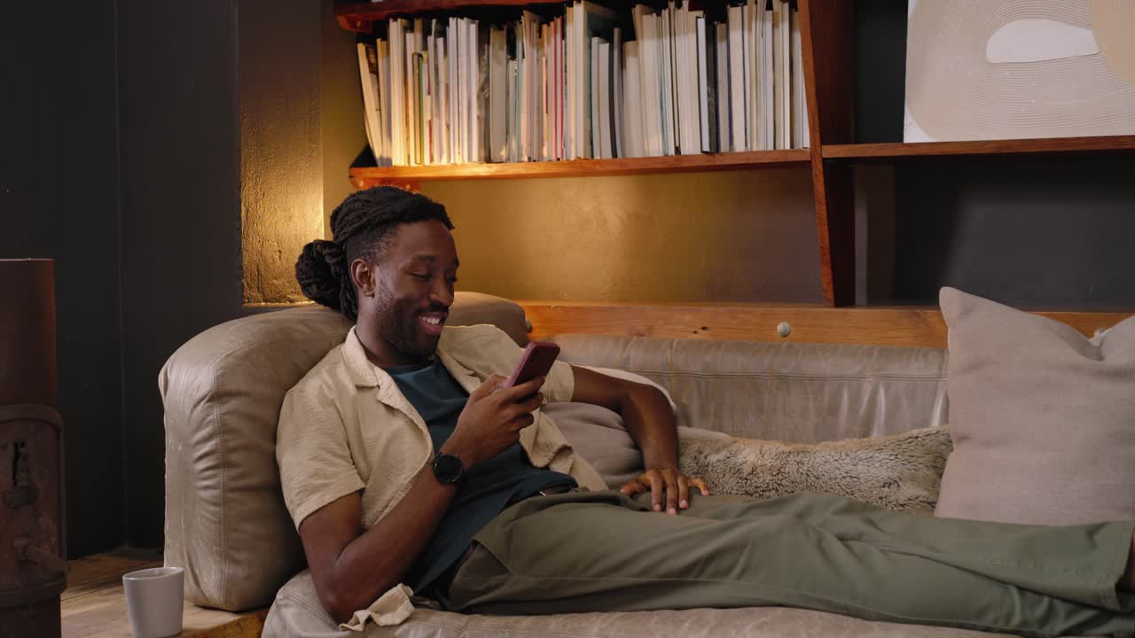 African man reclining on sofa at home, receiving smartphone alert, tapping and reacting to message
